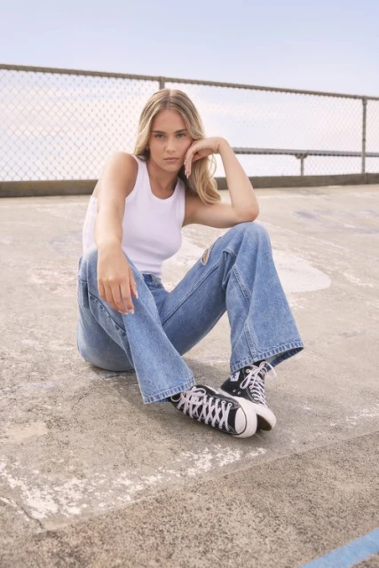 A blonde adult woman sits on a concrete surface wearing ripped blue jeans and a white tank top with black and white sneakers.