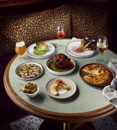 Round restaurant table laden with multiple gourmet dishes, grilled vegetables, a whole roasted cauliflower, and drinks.