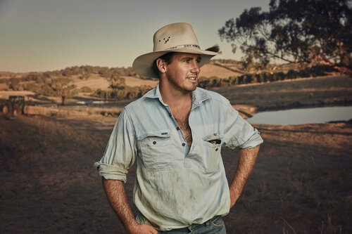 Outdoor portrait of a man wearing a hat and blue work shirt, standing on a farm with rolling hills and a small pond in the background.