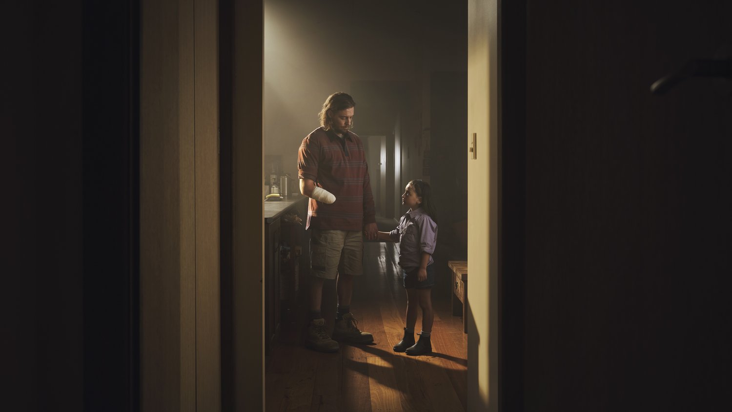 An adult and child are holding hands in a doorway, bathed in soft light. The adult wears a plaid shirt and the child looks up.
