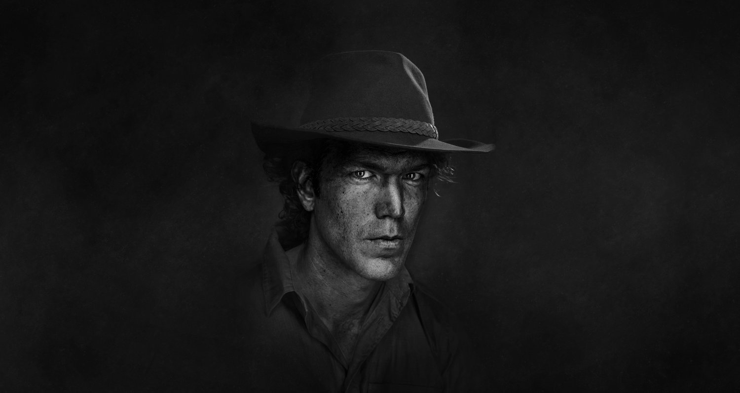 Monochrome portrait of a man with a hat looking at the camera against a dark background, creating a dramatic and intense image.