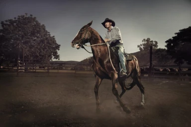 An older man wearing a cowboy hat and striped shirt rides a dark brown horse outdoors on a dusty dirt track.