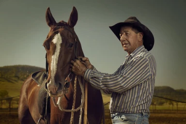 Elderly man in cowboy hat and striped shirt standing beside a brown horse outdoors in Melbourne by Tom Franks