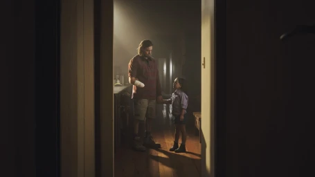 A man with a bandaged arm and a young girl standing indoors on a wooden floor in a dimly lit domestic setting.
