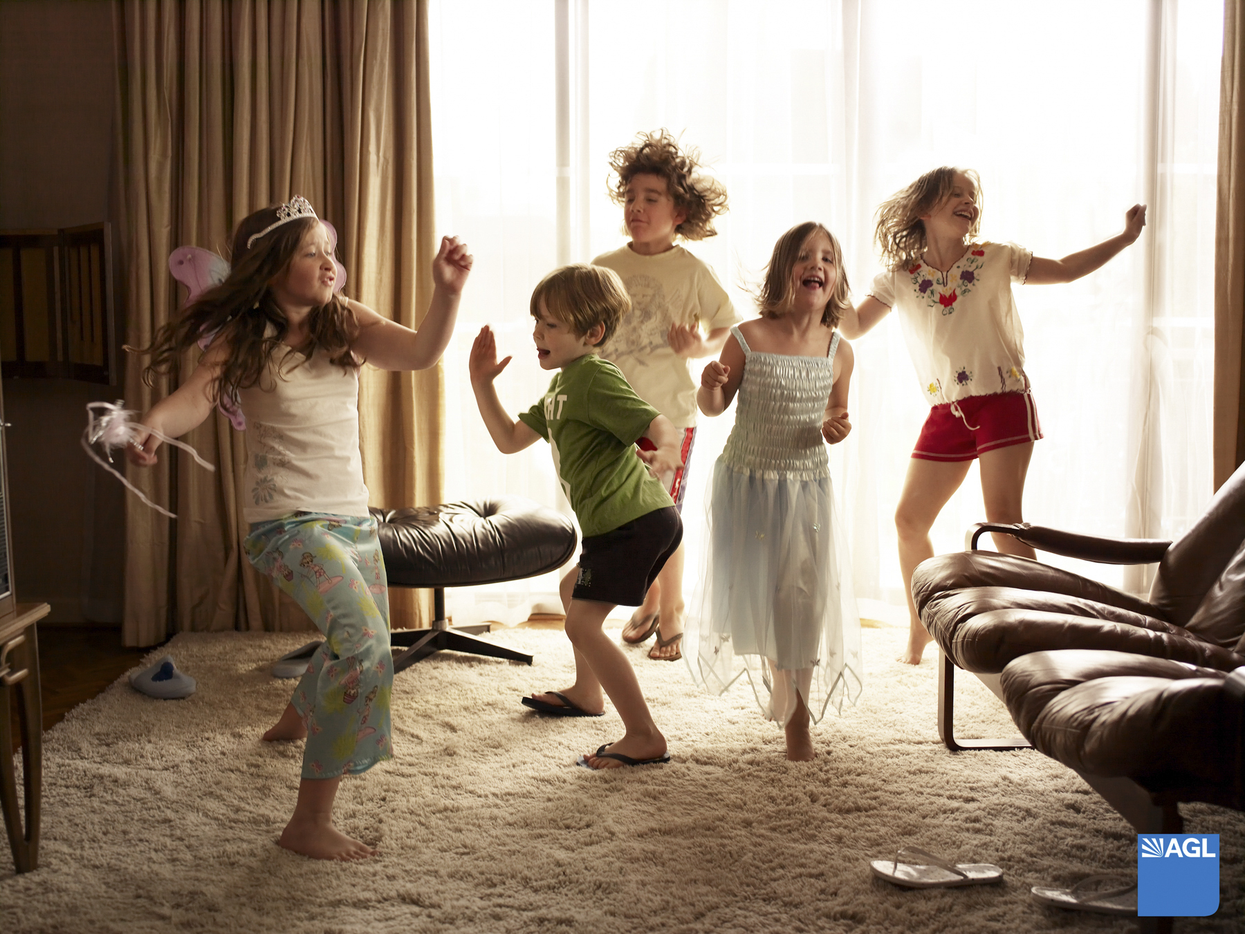 Five happy children dance and play in a brightly lit living room, with sunlight streaming through sheer curtains and an armchair in view.