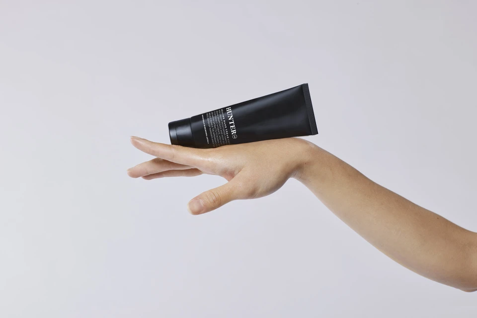 A black tube of Hunter Lab hand cream is held on a woman's hand against a white background, showcasing its sleek design.