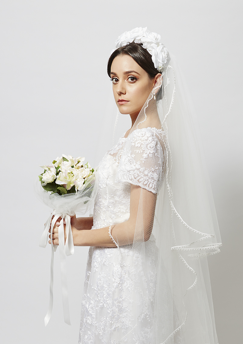 Portrait of a beautiful bride in a white lace wedding dress and veil holding a bouquet of flowers against a white background.
