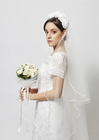 Portrait of a beautiful bride in a white lace wedding dress and veil holding a bouquet of flowers against a white background.