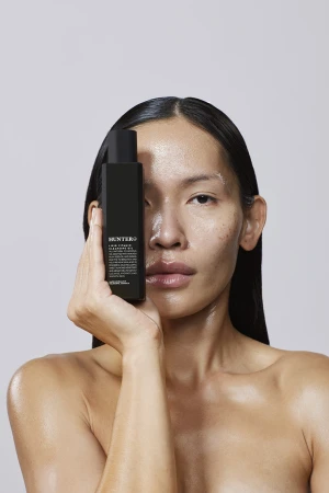 Adult female poses in studio with black bottle of cleanser. Beauty shot features moisturized skin, neutral background, clean aesthetic.