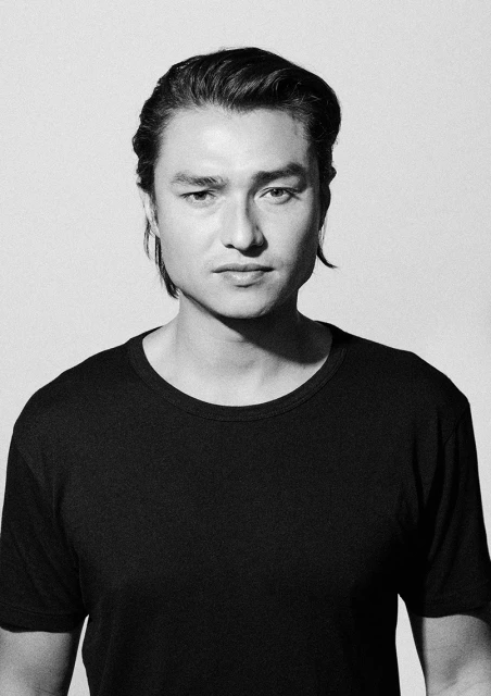 Black and white portrait of an adult male with dark hair wearing a T-shirt against a neutral background in a photo studio.