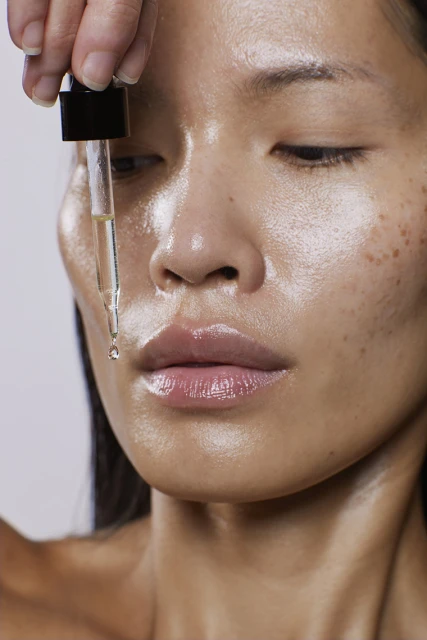 Close-up of an Asian woman's face with glossy skin and a hand applying facial serum via a dropper, against a light neutral background.