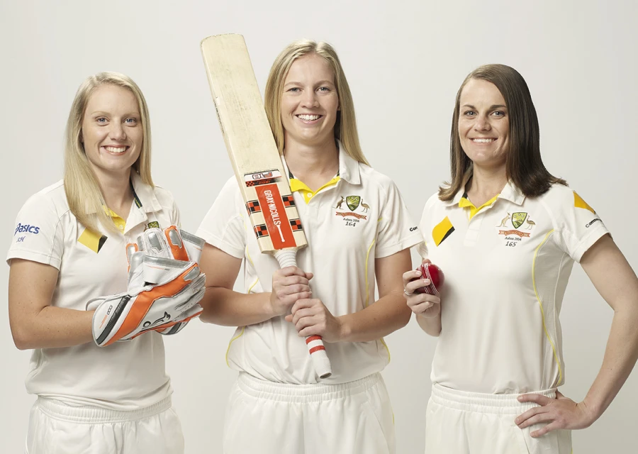 Three female cricket players pose in uniform with a bat, ball, and gloves against a white background, looking directly at the camera.