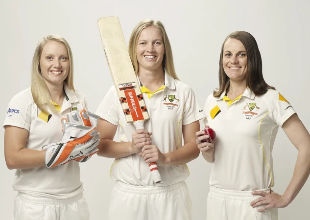 Three female cricket players pose in uniform with a bat, ball, and gloves against a white background, looking directly at the camera.
