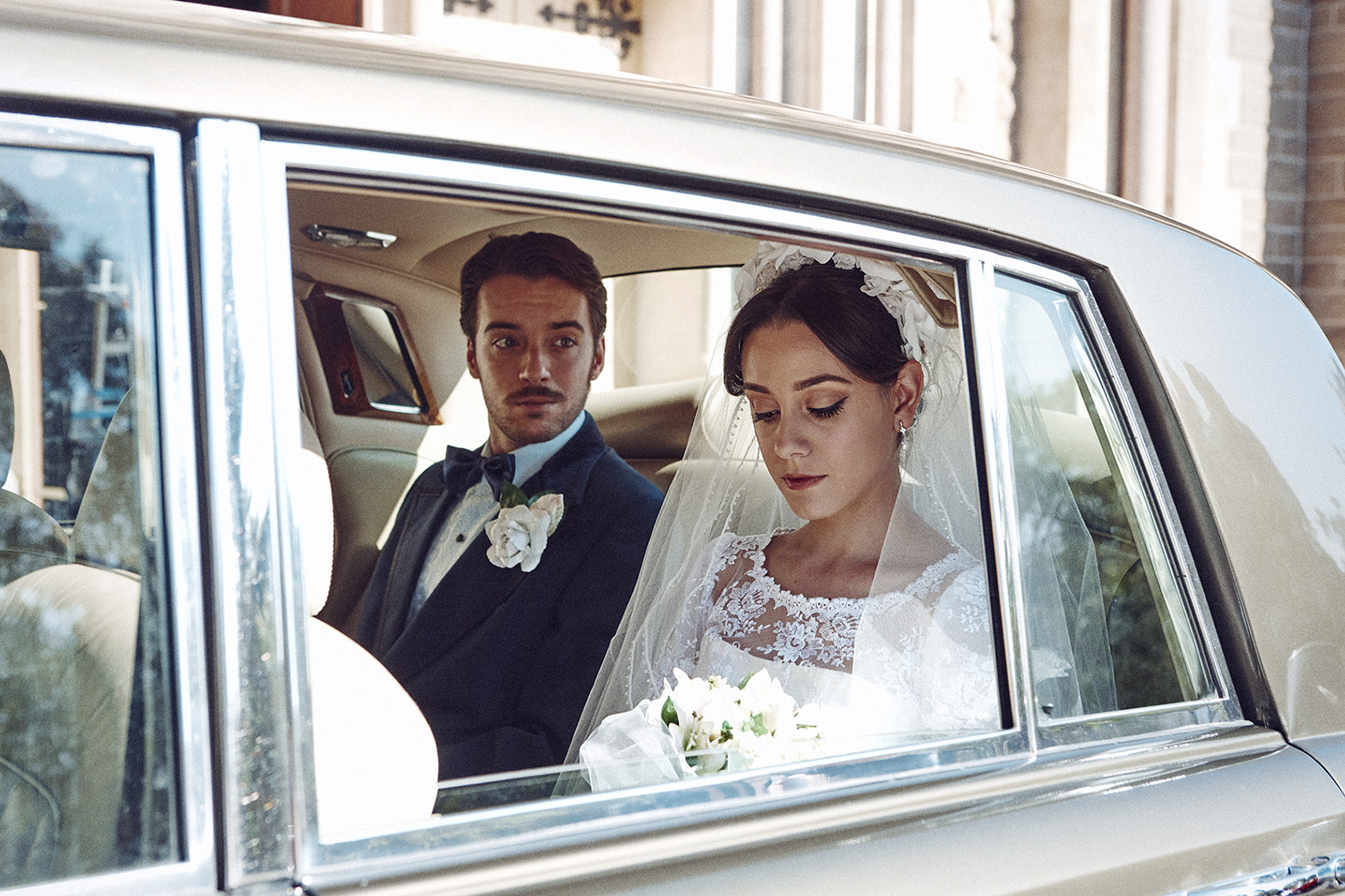 A bride and groom ride in the back seat of a classic car on their wedding day, with formal wear, white lace dress, and bow tie.