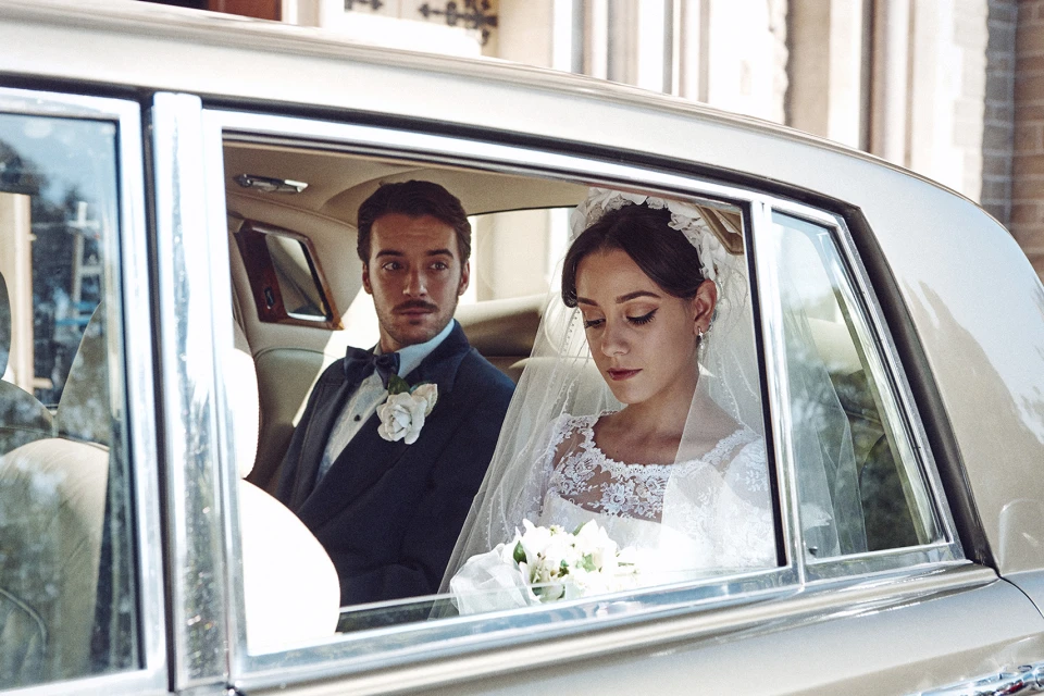 A bride and groom ride in the back seat of a classic car on their wedding day, with formal wear, white lace dress, and bow tie.