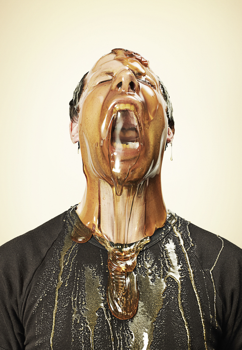 A person with honey pouring all over their face and black shirt. The individual's mouth is open, and the background is off-white.
