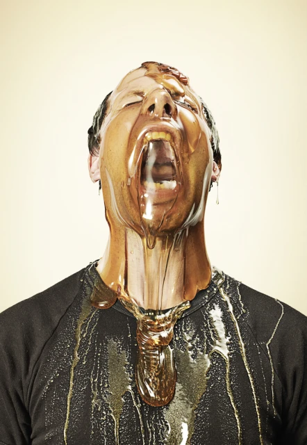 A person with honey pouring all over their face and black shirt. The individual's mouth is open, and the background is off-white.