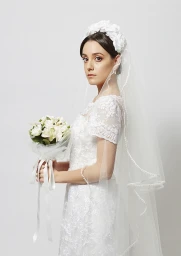 Bride in a white lace wedding dress with a floral veil holding a bouquet against a plain white background.