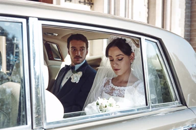 Bride in lace dress and groom in tuxedo seated inside a vintage car looking out the window