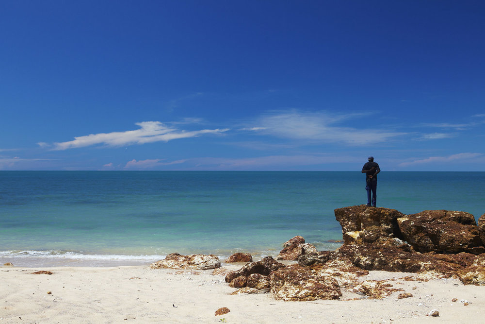 Adult male stands on rocks overlooking a beach and the ocean under a blue sky. The scene captures coastal serenity.