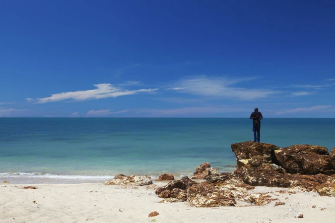 Lone person standing on coastal rocks overlooking a turquoise ocean under a bright blue sky, photographed in Melbourne.