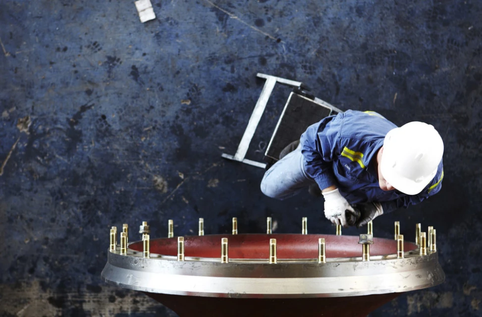 Overhead view of a worker in a hardhat and blue overalls tightening bolts on a large red and silver industrial component.