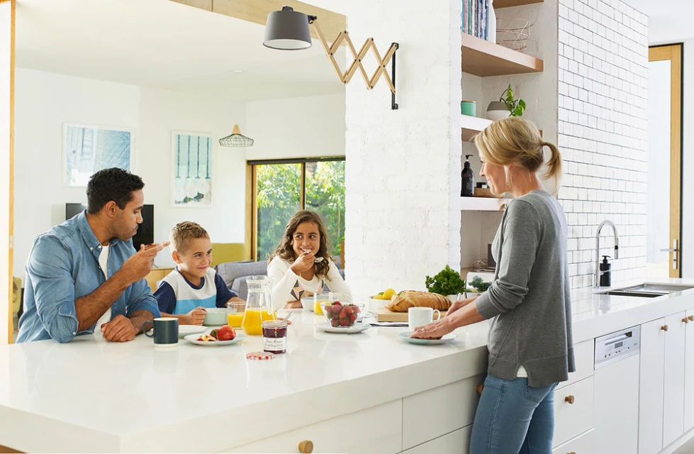 A family—father, mother, son, and daughter—sitting at a white kitchen island eating breakfast with juice, bread, and fruit.