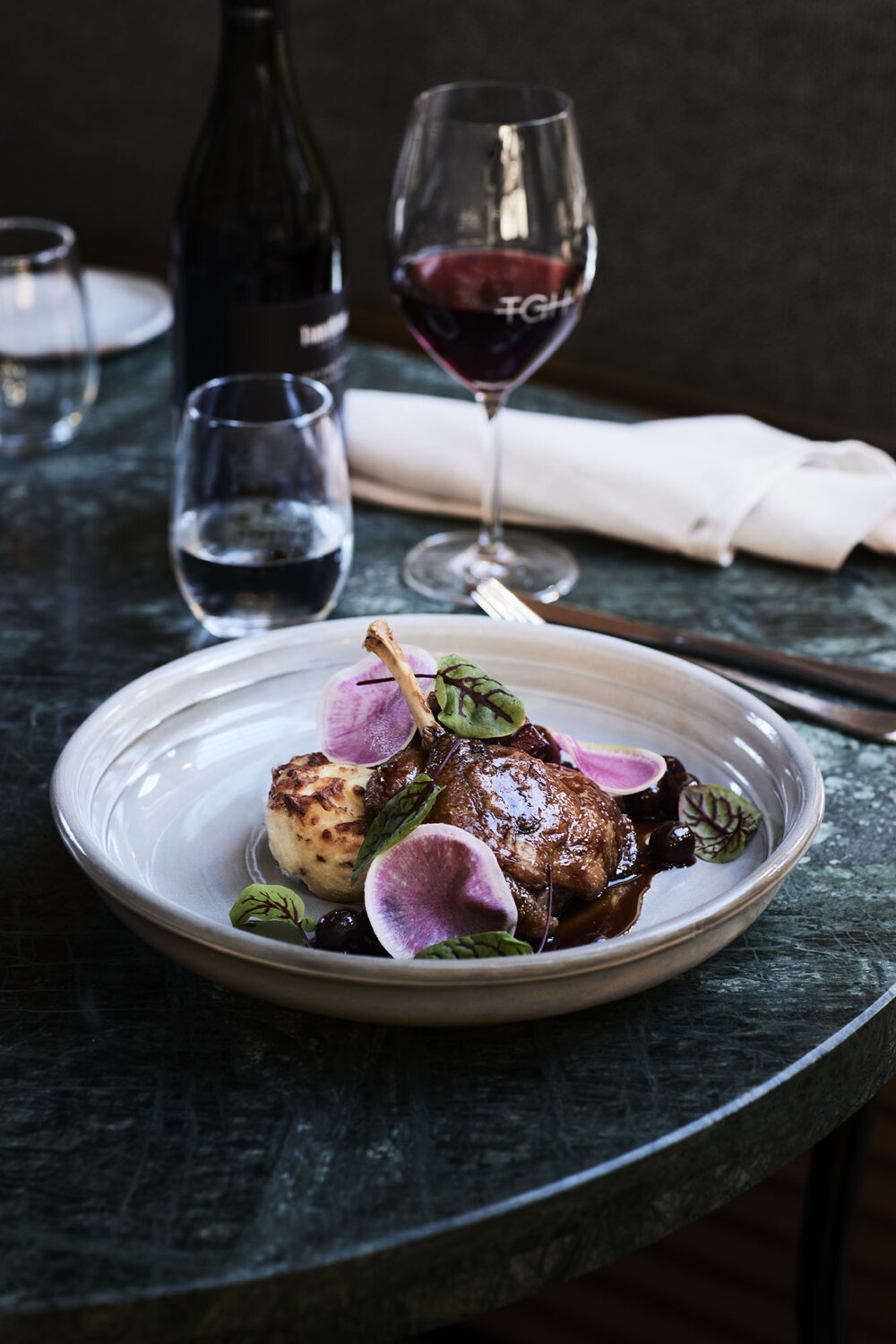 Overhead shot of duck confit plated with potato and purple radish alongside wine glasses and a bottle on a marble table.