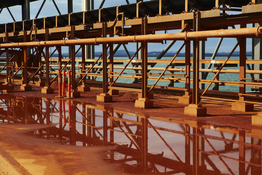 Industrial infrastructure featuring a network of metal beams and pipes, with orange and white safety bollards and a body of water visible in the background.