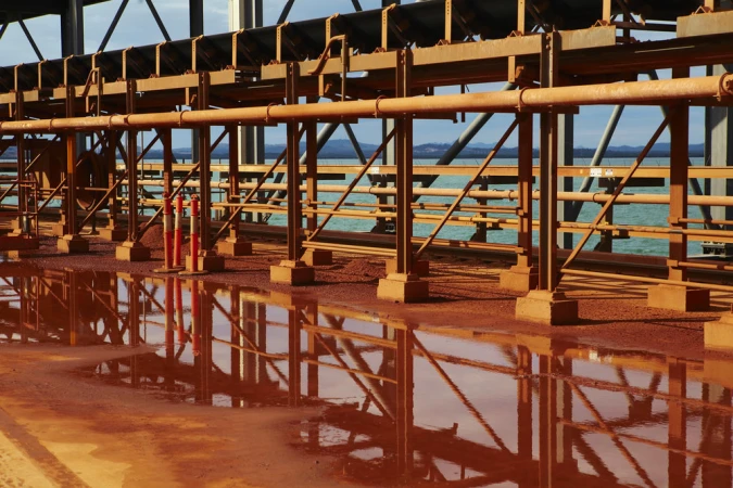 Industrial infrastructure featuring a network of metal beams and pipes, with orange and white safety bollards and a body of water visible in the background.