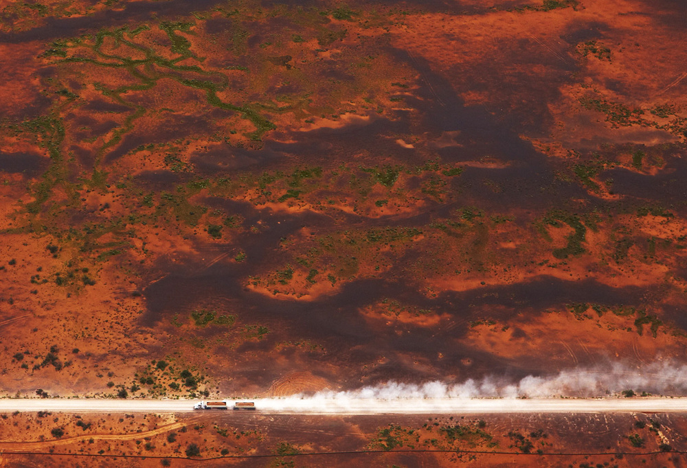Aerial shot of two semi-trucks driving on a dirt road in a desert landscape, kicking up dust. The ground is a reddish-brown color.