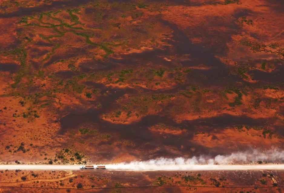 Aerial shot of two semi-trucks driving on a dirt road in a desert landscape, kicking up dust. The ground is a reddish-brown color.