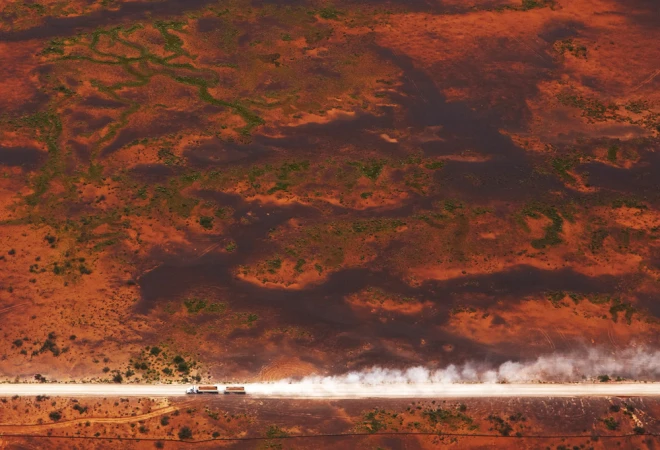 Aerial shot of two semi-trucks driving on a dirt road in a desert landscape, kicking up dust. The ground is a reddish-brown color.
