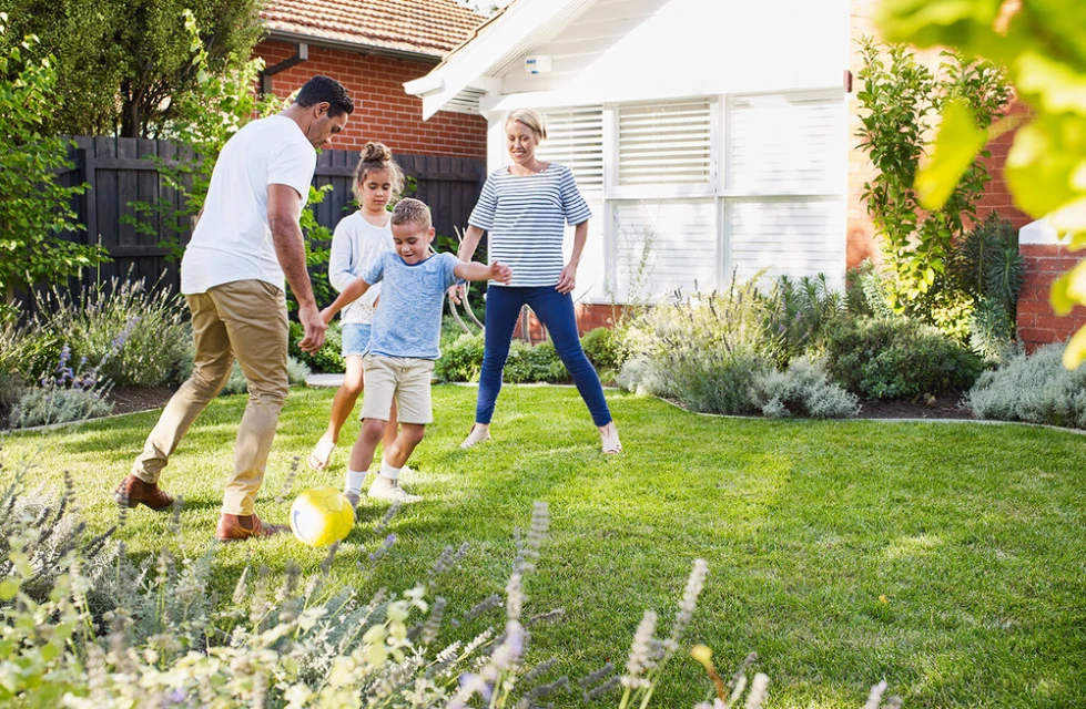 Family of four plays soccer in backyard on a sunny day. Father kicks the yellow ball to his son. Lush green grass, plants, and house.