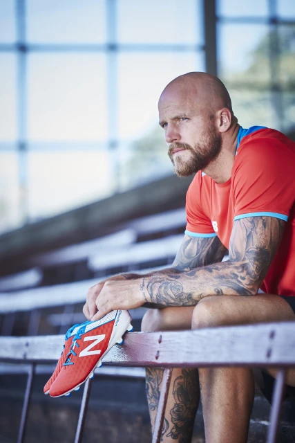 A bald man with a beard, wearing a red sports shirt, sits on stadium seating and holds an orange-and-white New Balance soccer shoe.