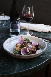 Plated roasted duck dish with radish slices in a ceramic bowl on a dark marble table with wine glasses.