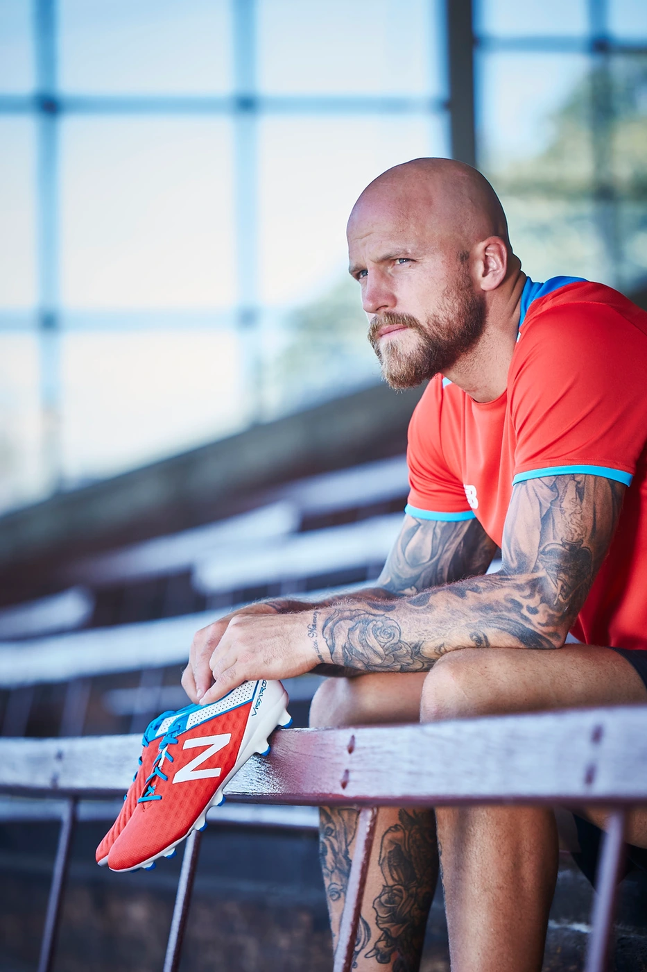 Bald tattooed man seated on wooden bleachers holding a red soccer cleat in his hands