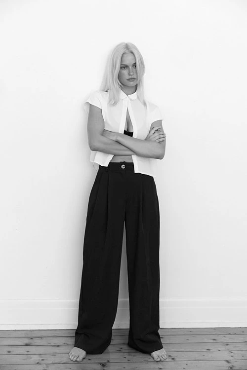 Blonde woman with arms crossed wearing a white shirt and black trousers against a plain white wall by Maddie Roux