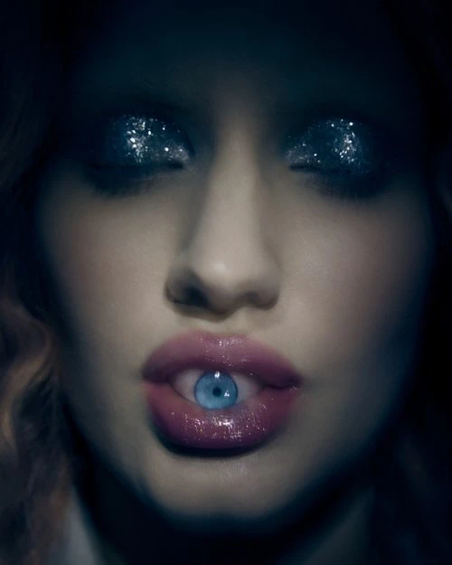 Close-up of a woman's face with an eye photoshopped in her mouth, glitter eye makeup, and pink lipstick, set against a dark background.