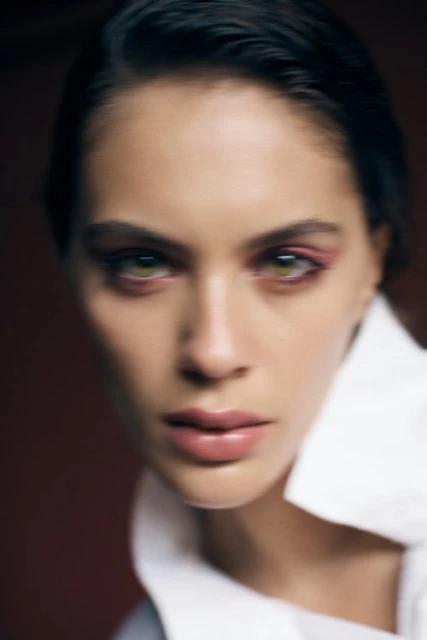 Close-up portrait of an adult woman with green eyes and pink eyeshadow, wearing a white collared shirt in a studio setting.