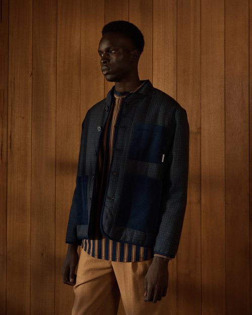 An adult male in a dark blue jacket and tan pants poses against a wood wall. The clothing is a striped shirt and textured jacket.