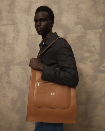 Black man holding a large brown leather tote bag wearing a dark jacket against a textured concrete wall