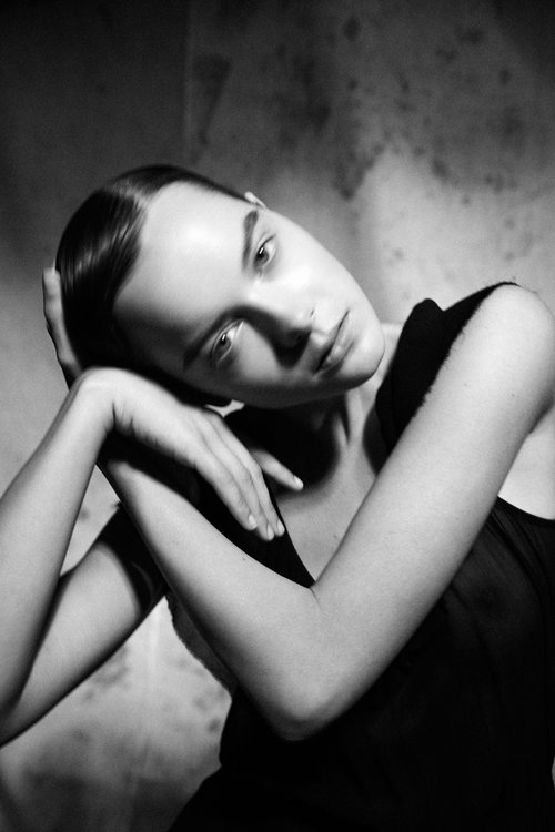 Monochromatic portrait of a woman with dark hair resting her head on her hands against a textured backdrop in a close-up shot.