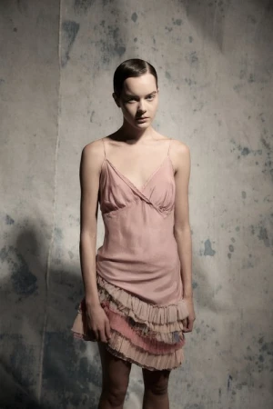 Model with slicked-back hair wearing a pink slip dress with layered ruffles against a mottled backdrop