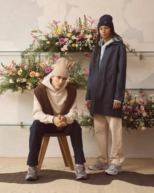 Two people, one seated and one standing, model clothing in front of a floral display, photographed indoors with soft, diffused lighting.