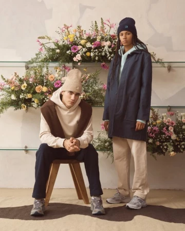 Two people, one seated and one standing, model clothing in front of a floral display, photographed indoors with soft, diffused lighting.