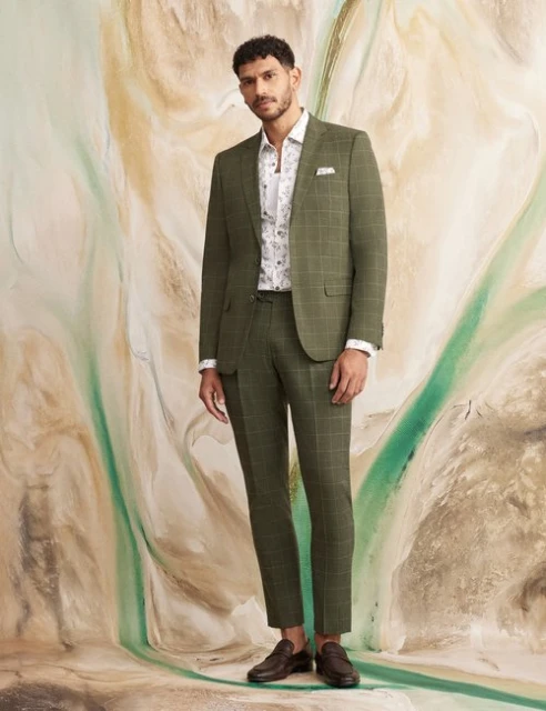 Man in a green plaid suit, patterned shirt, and loafers poses against a painted backdrop in a full-length studio shot, looking at the camera.