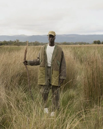 Black man standing in tall grass wearing a quilted olive coat and cap while holding a wooden staff.