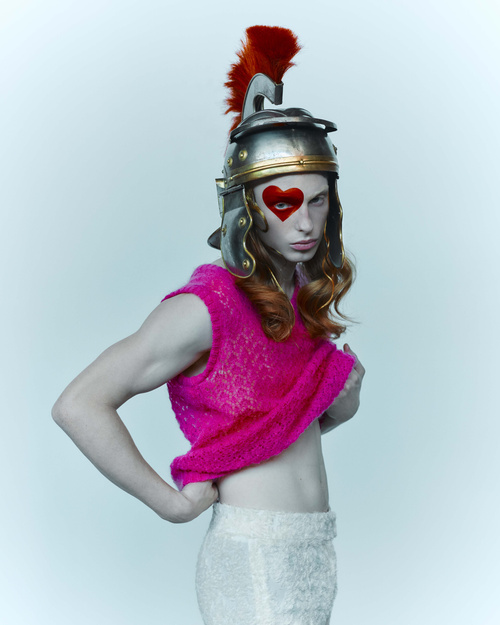 Person in a pink tank top, white skirt, and silver Roman gladiator helmet with a heart painted on face posing in a photo studio.