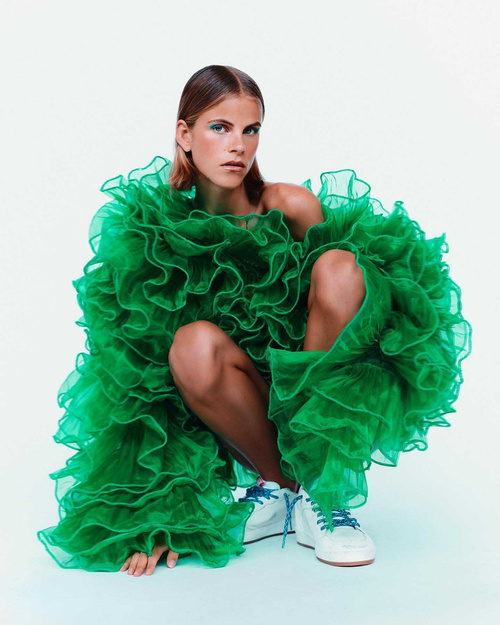 A woman with light brown hair poses in a squatting position wearing a bright green ruffled dress and white sneakers on a white backdrop.